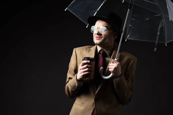 Homem elegante confuso em terno bege, chapéu e óculos segurando guarda-chuva e café para ir isolado em preto — Fotografia de Stock