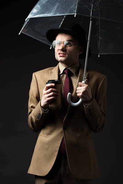 Confus homme élégant en costume beige, chapeau et lunettes tenant parapluie et café pour aller isolé sur noir — Photo de stock