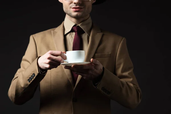 Vista recortada de hombre elegante en traje beige sosteniendo taza de café y platillo aislado en negro - foto de stock