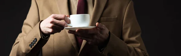 Vista recortada de hombre elegante en traje beige sosteniendo taza de café y platillo aislado en negro, plano panorámico - foto de stock