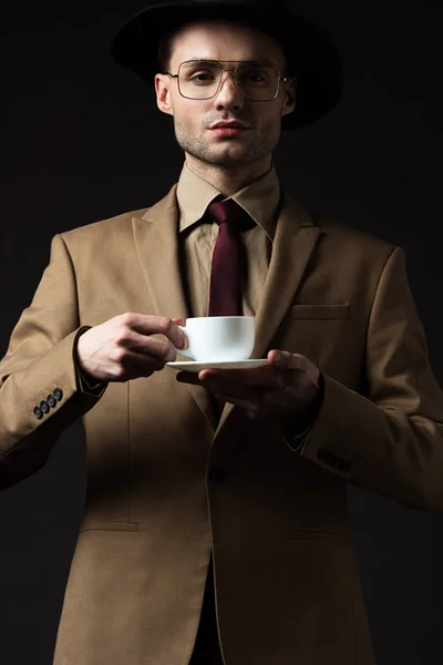 Elegante hombre en traje beige sosteniendo taza de café y platillo aislado en negro - foto de stock