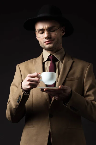 Hombre elegante en traje beige, sombrero y gafas con taza de café y platillo aislado en negro - foto de stock