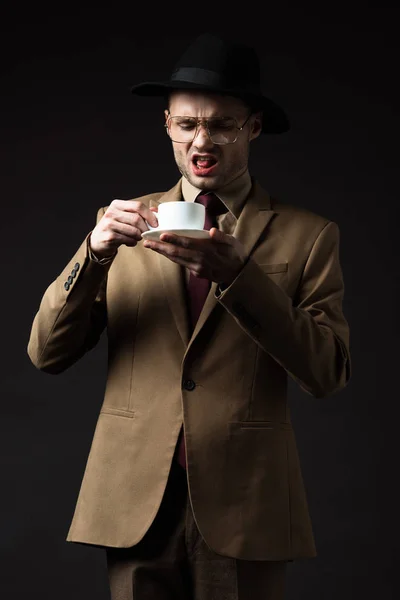 Displeased elegant man in beige suit, hat and eyeglasses holding coffee cup and saucer isolated on black — Stock Photo