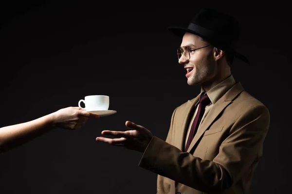 Lächelnder eleganter Mann in beigem Anzug, Hut und Brille, der Kaffee vom Kellner nimmt, isoliert auf schwarz — Stockfoto