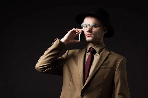 Hombre serio elegante en traje beige, sombrero y gafas que hablan en el teléfono inteligente aislado en negro - foto de stock