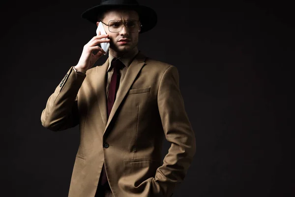 Serio uomo elegante in abito beige, cappello e occhiali che parla su smartphone isolato su nero — Foto stock