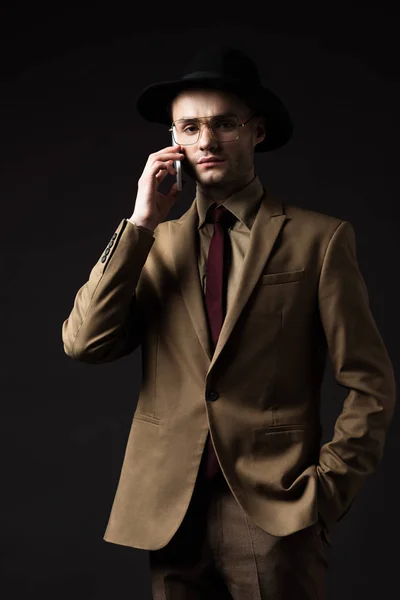 Serious elegant man in beige suit, hat and eyeglasses talking on smartphone isolated on black — Stock Photo