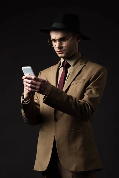 Серьезный элегантный мужчина в бежевом костюме, шляпе и очках, используя смартфон, изолированный на черном — стоковое фото