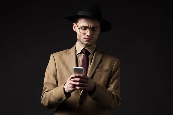 Serio uomo elegante in abito beige, cappello e occhiali da vista utilizzando smartphone isolato su nero — Foto stock