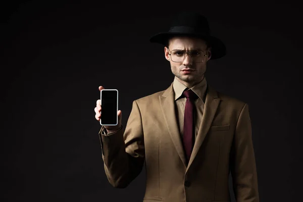 Homme élégant sérieux en costume beige, chapeau et lunettes tenant smartphone avec écran blanc isolé sur noir — Photo de stock