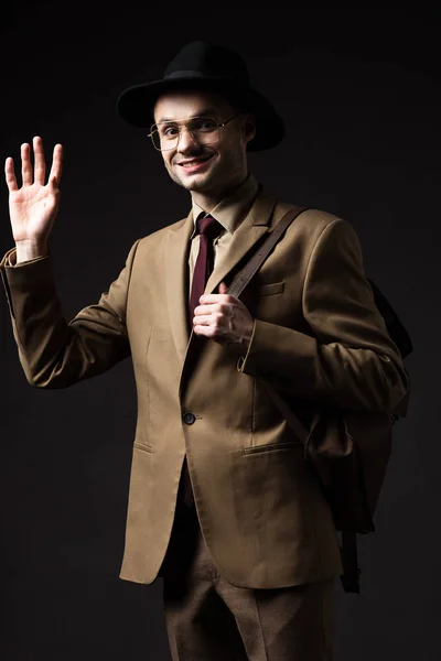 Smiling elegant man in beige suit, hat and eyeglasses with brown leather backpack waving hand isolated on black — Stock Photo