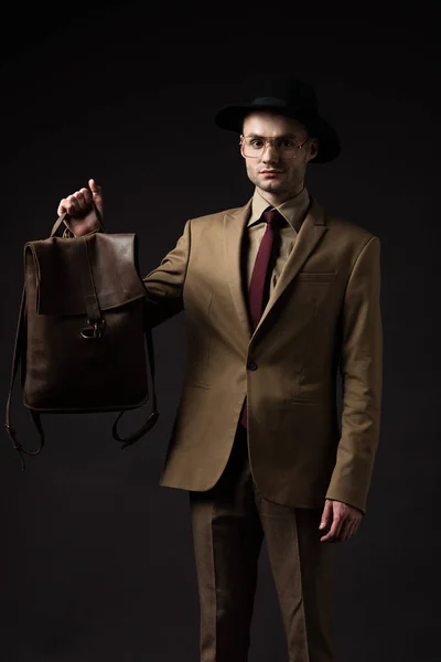 Élégant homme en costume beige, chapeau et lunettes tenant sac à dos en cuir brun isolé sur noir — Photo de stock