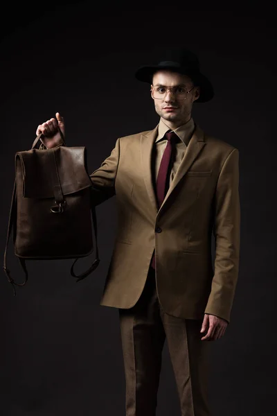 Elegante hombre en traje beige, sombrero y gafas con mochila de cuero marrón aislada en negro - foto de stock