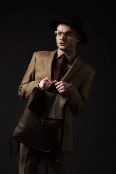 Hombre elegante preocupado en traje beige, sombrero y gafas con mochila de cuero marrón aislado en negro - foto de stock