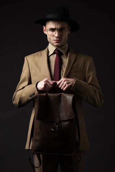 Hombre elegante y tenso en traje beige, sombrero y gafas con mochila de cuero marrón aislada en negro - foto de stock