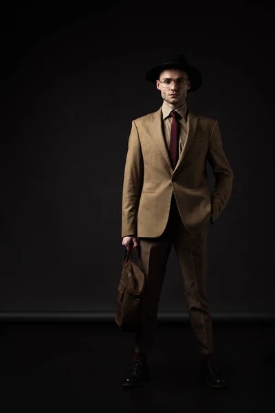 Élégant homme en costume beige, chapeau et lunettes tenant sac en cuir marron avec la main dans la poche sur noir — Photo de stock