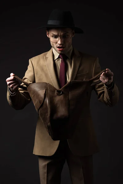 Homme élégant choqué en costume beige, chapeau et lunettes à la recherche dans un sac en cuir brun isolé sur noir — Photo de stock
