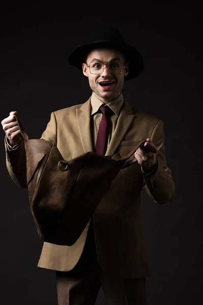 Щасливий елегантний чоловік у бежевому костюмі, капелюсі та окулярах, дивлячись у коричневий шкіряний мішок ізольований на чорному — стокове фото