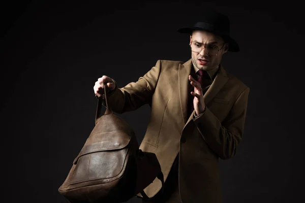 Stressed elegant man in beige suit, hat and eyeglasses looking in brown leather bag isolated on black — Stock Photo