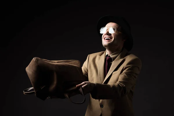 Smiling elegant man in beige suit, hat and eyeglasses shaking out brown leather bag isolated on black — Stock Photo