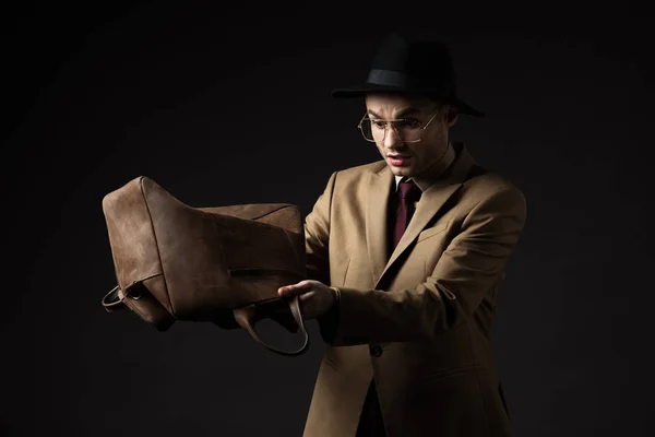 Hombre elegante acentuado en traje beige, sombrero y gafas que sacuden el bolso de cuero marrón aislado en negro - foto de stock