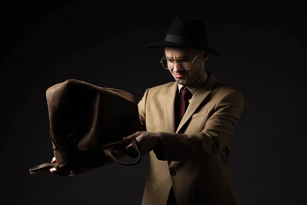 Triste hombre elegante en traje beige, sombrero y anteojos sacuden bolsa de cuero marrón aislado en negro — Stock Photo