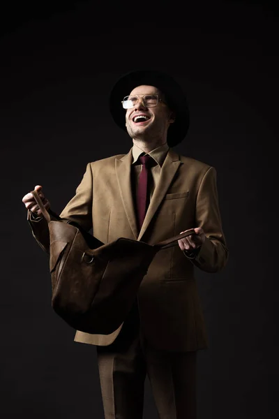 Hombre elegante feliz en traje beige, sombrero y gafas con bolsa de cuero marrón aislada en negro - foto de stock