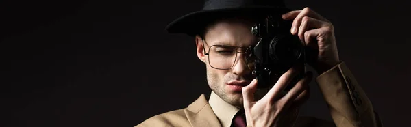 Elegante hombre en traje beige, sombrero y anteojos tomando fotos en cámara de cine aislado en negro, plano panorámico - foto de stock