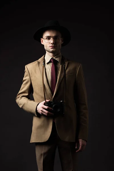 Hombre elegante en traje beige, sombrero y gafas con cámara de película aislada en negro - foto de stock