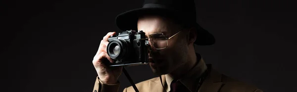 Elegante hombre en traje beige, sombrero y anteojos tomando fotos en cámara de cine aislado en negro, plano panorámico - foto de stock