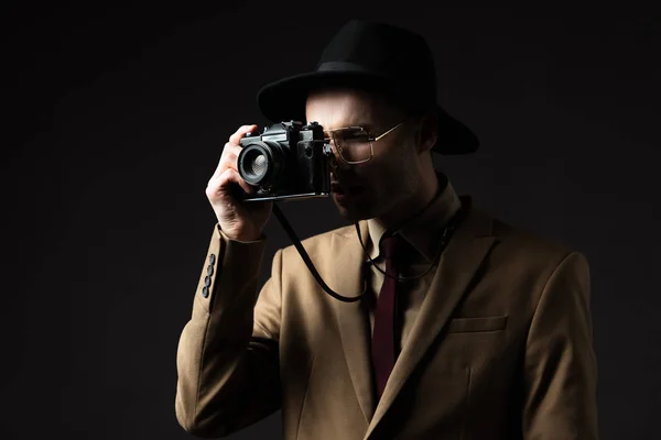 Elegant man in beige suit, hat and eyeglasses taking picture on film camera isolated on black — Stock Photo