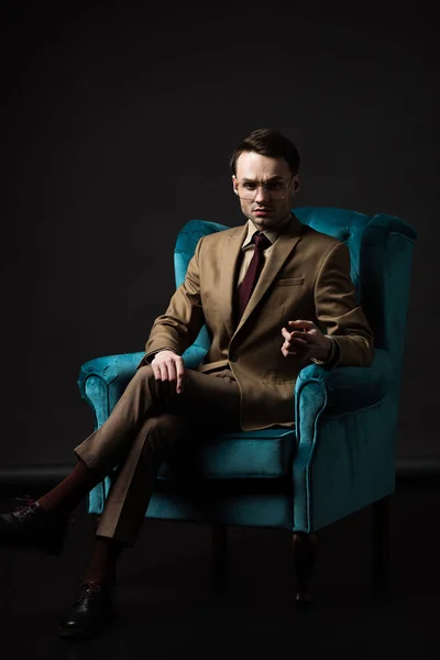 Серьезный элегантный мужчина в очках в бежевом костюме сидит в синем велюровом кресле и указывает пальцем на камеру, изолированную на черном — стоковое фото