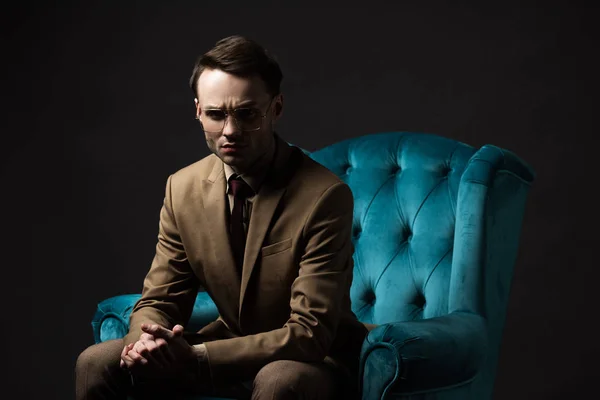 Élégant homme tendu en costume beige lunettes assis avec les mains serrées dans un fauteuil en velours bleu isolé sur noir — Photo de stock