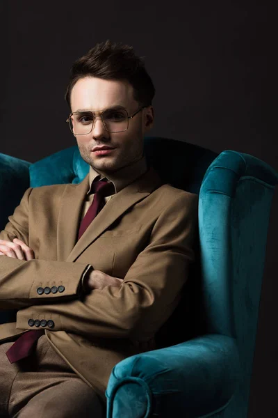 Elegant man in beige suit eyeglasses sitting in blue velour armchair with crossed arms isolated on black — Stock Photo