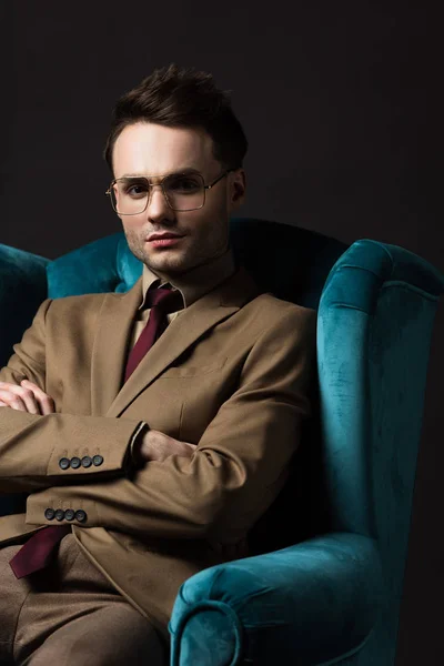 Elegante hombre en gafas de traje beige sentado en sillón de terciopelo azul con brazos cruzados aislados en negro - foto de stock