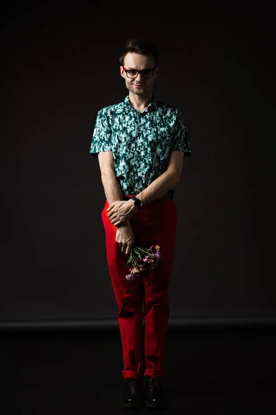 Shy fashionable man in eyeglasses in blue colorful shirt and red pants holding floral bouquet on black — Stock Photo