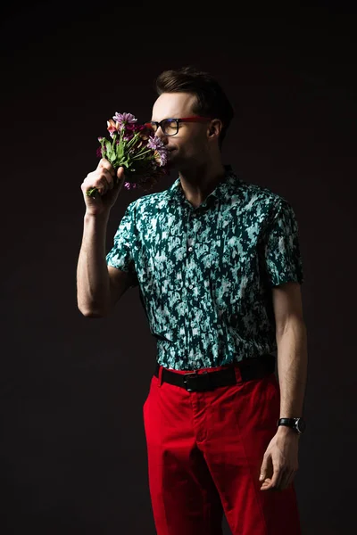 Fashionable man in eyeglasses in blue colorful shirt and red pants smelling floral bouquet isolated on black — Stock Photo