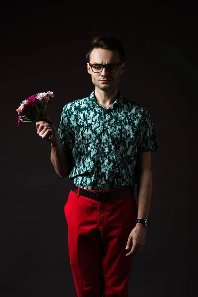 Uomo di moda sconvolto in occhiali in camicia colorata blu e pantaloni rossi tenendo bouquet floreale isolato su nero — Foto stock