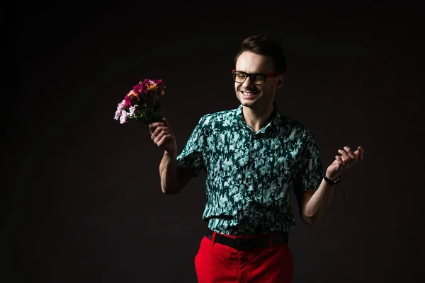 Щасливий модний чоловік в окулярах у синій барвистій сорочці і червоних штанях тримає квітковий букет ізольовано на чорному — стокове фото
