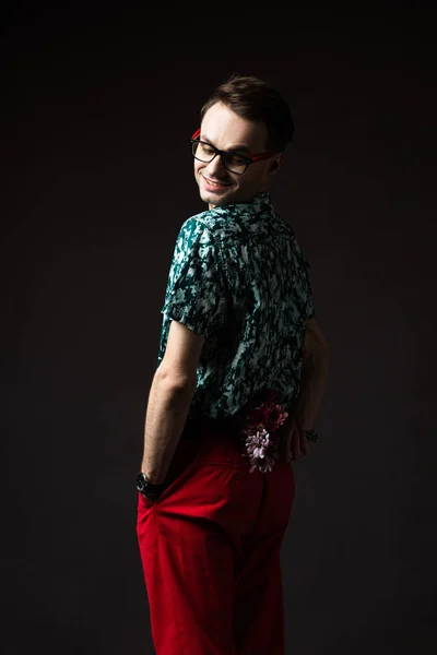 Smiling fashionable man in eyeglasses in blue colorful shirt and red pants holding floral bouquet behind back isolated on black — Stock Photo