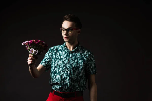 Surprised fashionable man in eyeglasses in blue colorful shirt and red pants holding floral bouquet isolated on black — Stock Photo
