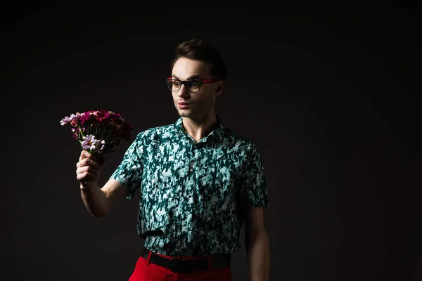 Homem elegante surpreso em óculos em camisa colorida azul e calças vermelhas segurando buquê floral isolado em preto — Fotografia de Stock