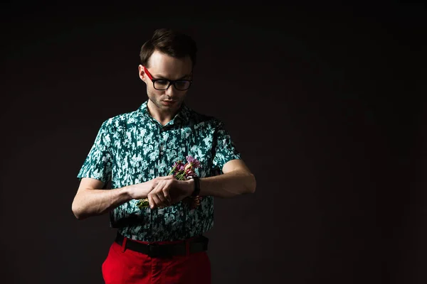 Модный мужчина в очках в синей красочной рубашке и красных брюках с цветочным букетом и проверкой времени, изолированного на черном — стоковое фото