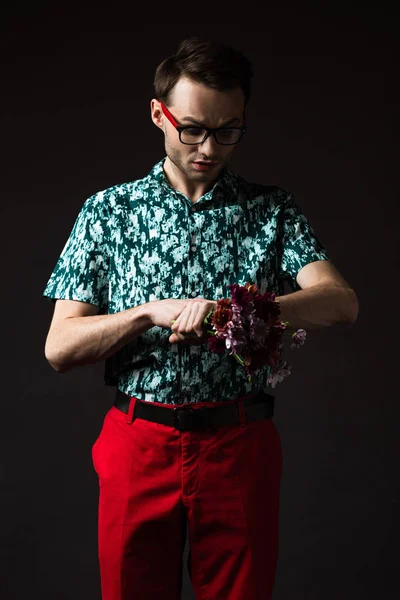 Fashionable man in eyeglasses in blue colorful shirt and red pants holding floral bouquet and checking time isolated on black — Stock Photo