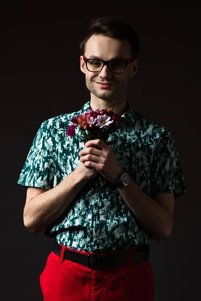 Smiling fashionable man in eyeglasses in blue colorful shirt and red pants holding floral bouquet isolated on black — Stock Photo