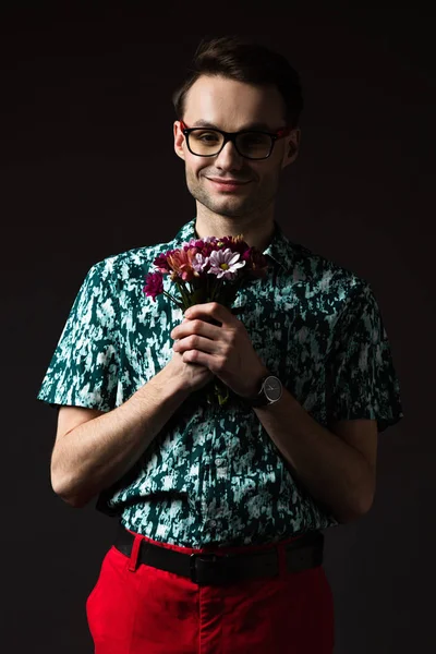 Lächelnder modischer Mann mit Brille in blauem, buntem Hemd und roter Hose mit Blumenstrauß auf schwarzem Hintergrund — Stockfoto