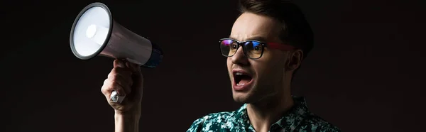 Fashionable man in eyeglasses in blue colorful shirt shouting in loudspeaker isolated on black, panoramic shot — Stock Photo