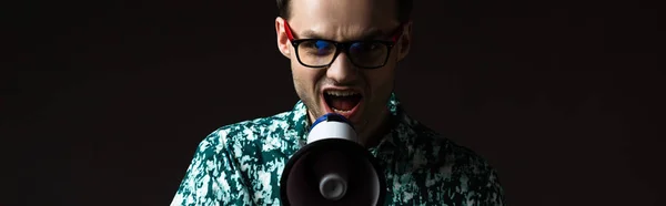 Fashionable man in eyeglasses in blue colorful shirt shouting in loudspeaker isolated on black, panoramic shot — Stock Photo