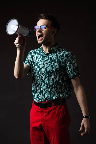 Fashionable man in eyeglasses in blue colorful shirt and red pants shouting in loudspeaker isolated on black — Stock Photo