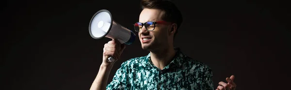 Fashionable man in eyeglasses in blue colorful shirt shouting in loudspeaker isolated on black, panoramic shot — Stock Photo